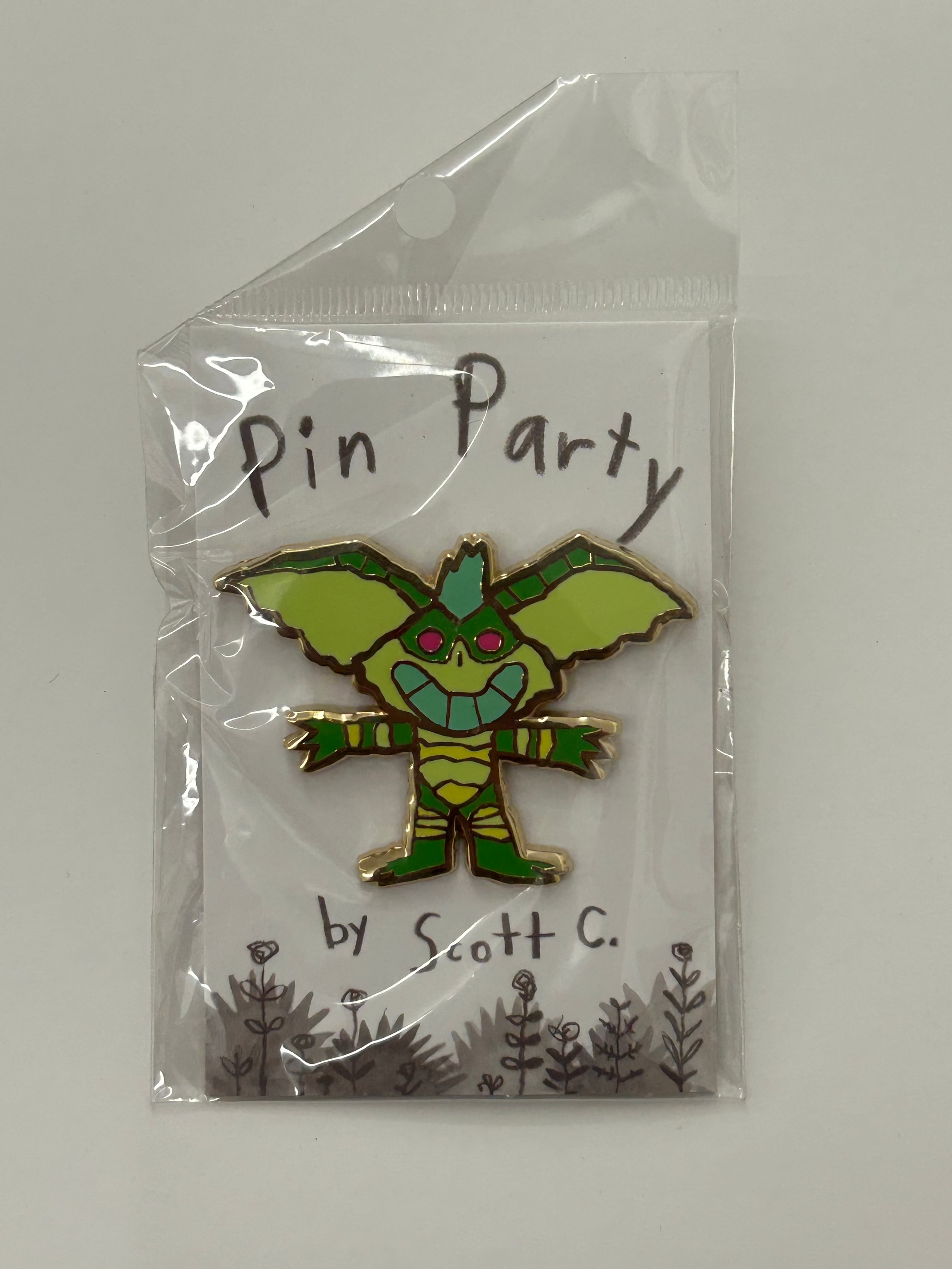 A green and yellow cartoon dragon hard enamel pin from Scott C. is shown on a Pin Party backing card with simple plant designs, packaged in a clear plastic bag.