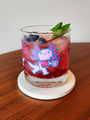 A pink iced drink garnished with blueberries and mint sits in a Spoke Art rocks glass featuring cheerful axolotl art, placed on a white coaster on a wooden surface.