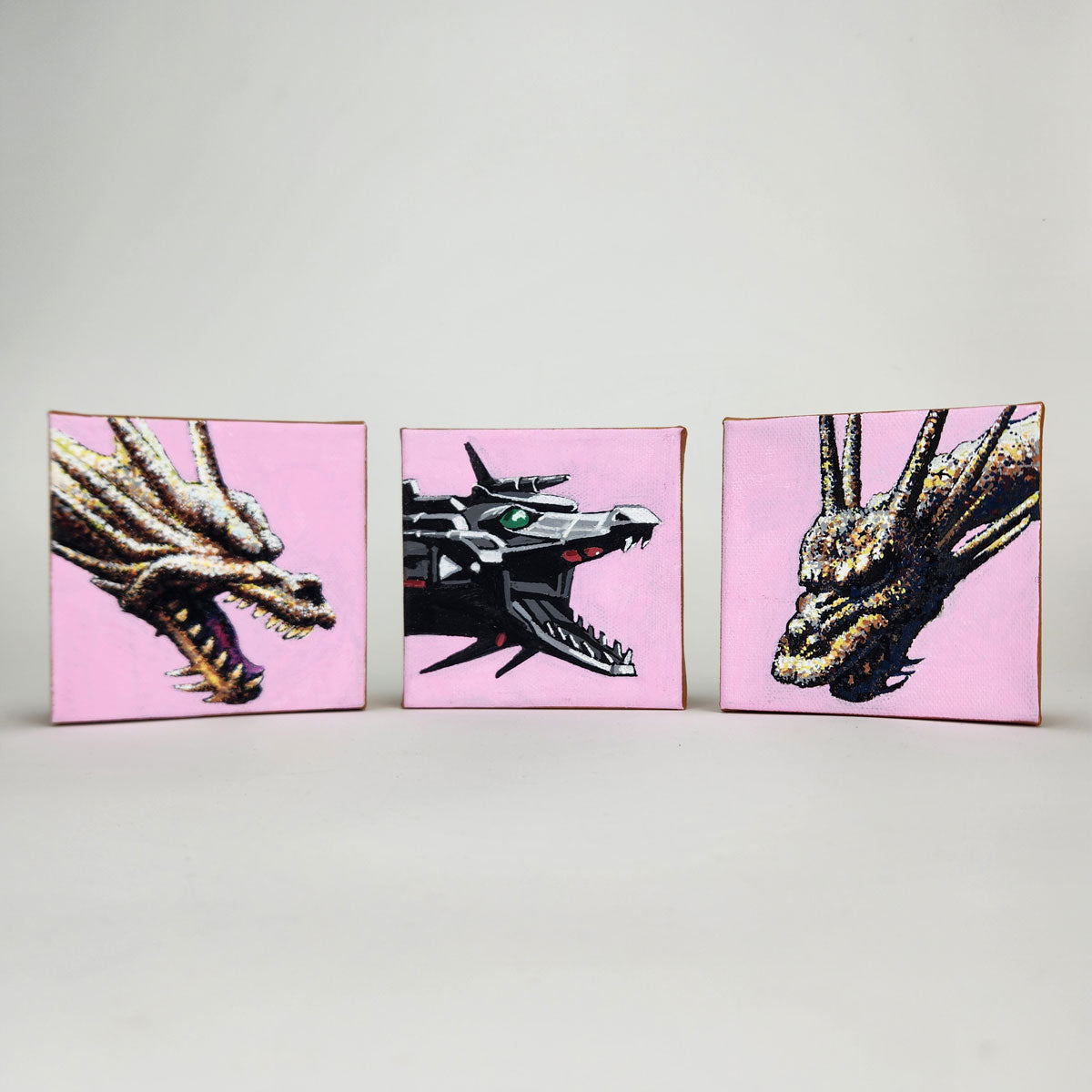 Three acrylic-on-canvas square pieces for Spoke Art’s 15th Anniversary Show feature pixel art dragon heads—a metallic, Mecha King Ghidorah-inspired design between two fierce dragons—set against pale pink backgrounds.