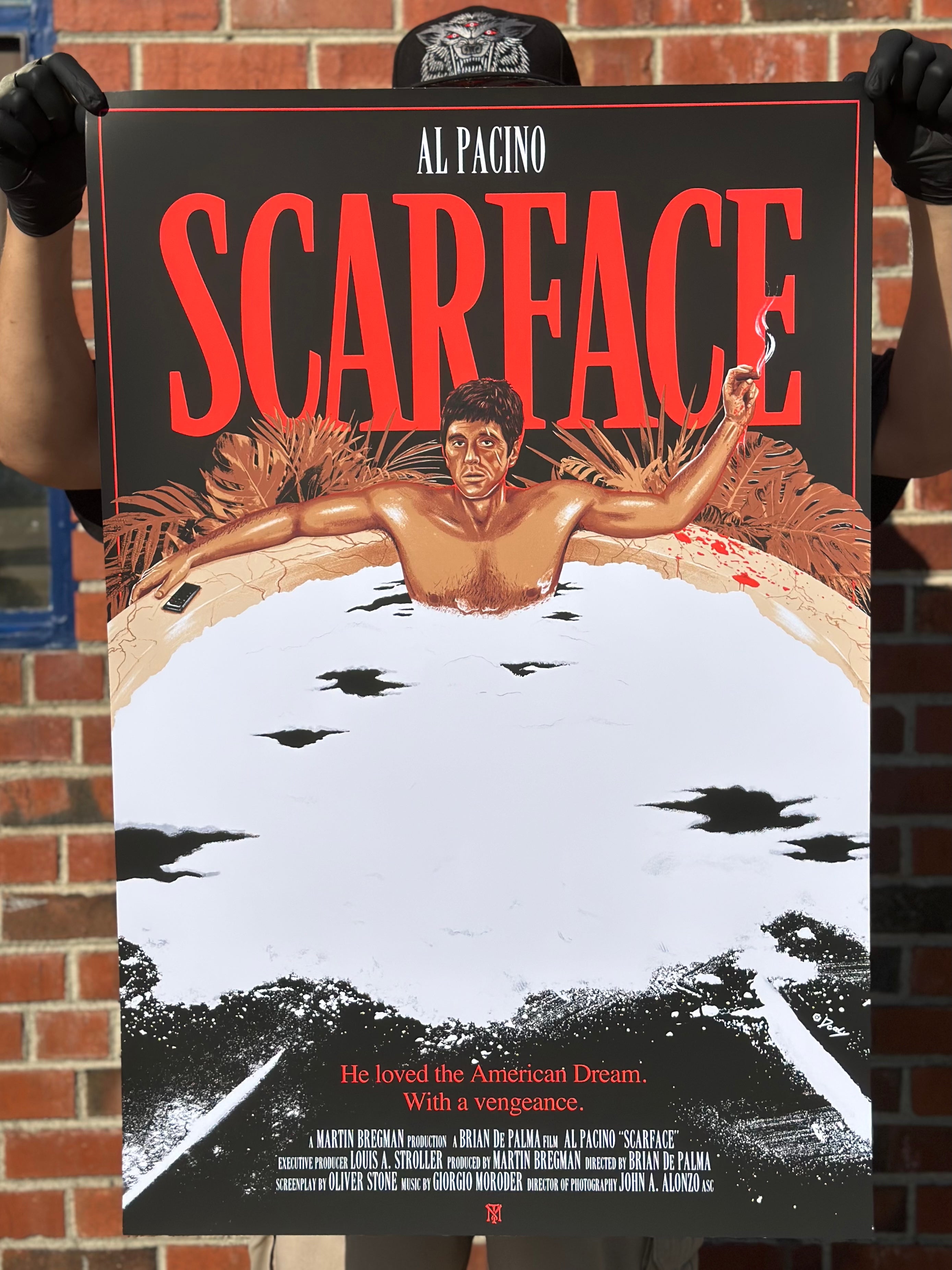 A person holds a limited edition Doaly Scarface screen print against a brick wall, featuring a man in a white suit with a gun, framed by bold red Scarface text. It highlights Al Pacino and includes an evocative American Dream tagline.