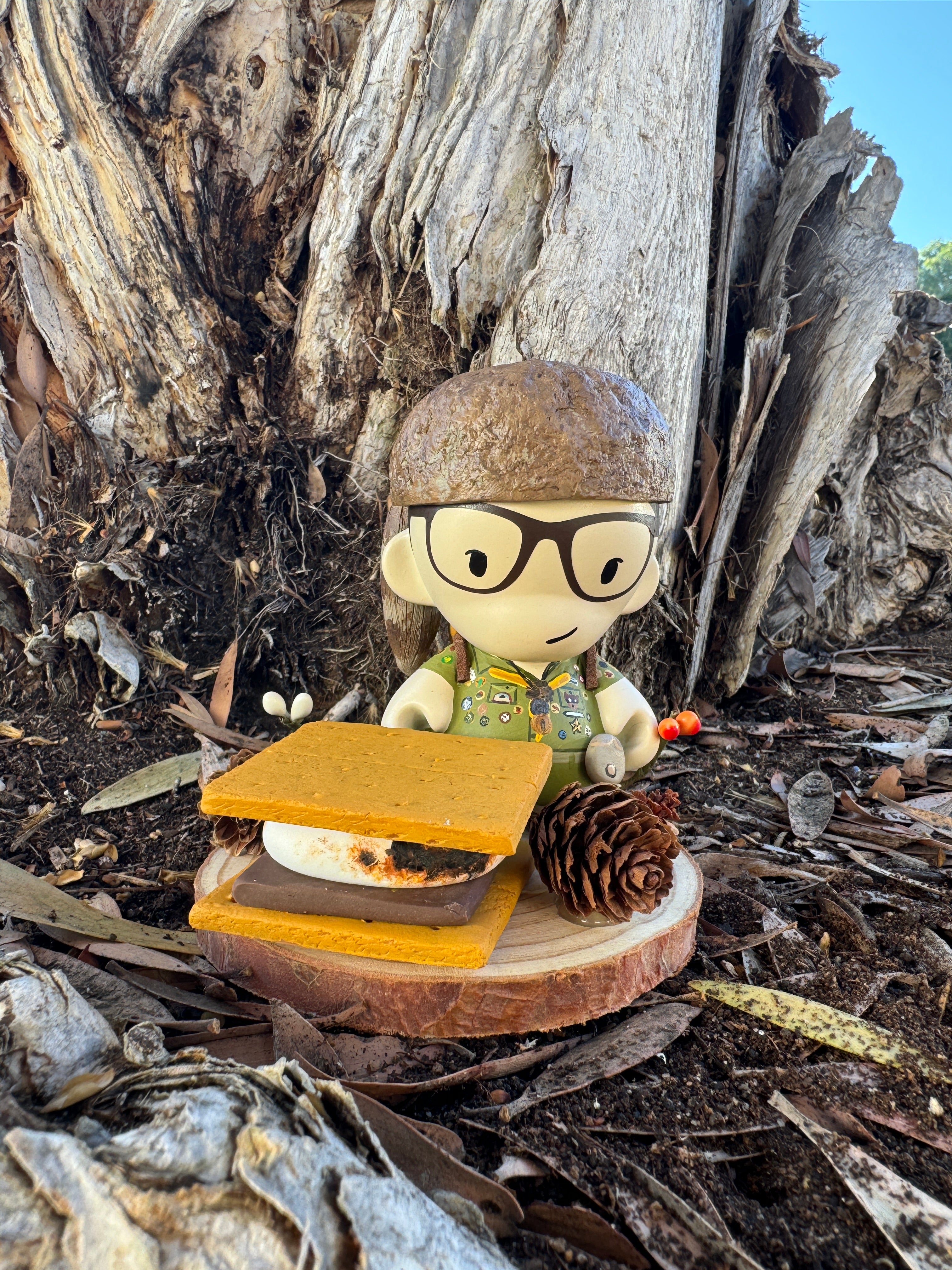 A Zard Apuya vinyl toy figure, wearing glasses and a green outfit with patches, stands next to a giant smore and pine cone. This scene, set against a textured tree trunk backdrop, captures the artful whimsy of Bad Dads IX.