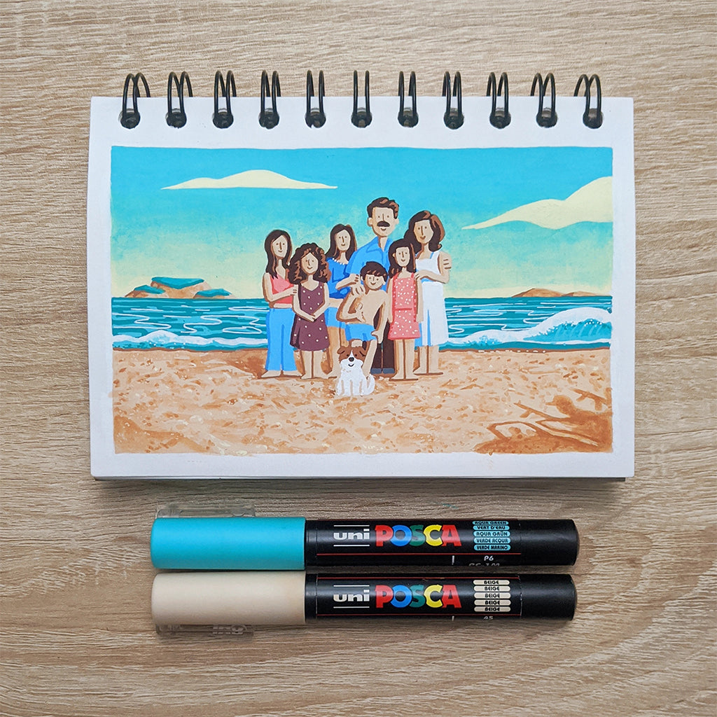 A James Chapman spiral-bound sketchbook showcases Im Still Here, featuring a family and their dog at the beach. Below it are two Posca markers in blue and beige, set against a backdrop of vibrant ocean and rugged rocks.