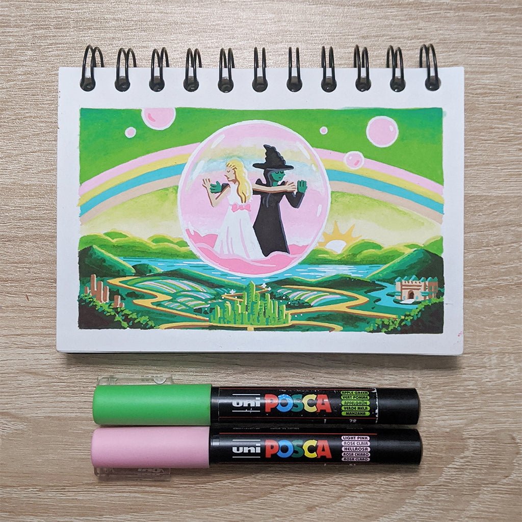 An illustration on a James Chapman spiral-bound notebook features a pink circle with two silhouetted figures: one in a white dress, the other in a witchs hat. Vibrant rainbows enhance the Wicked 2024 theme, with two marker pens below.