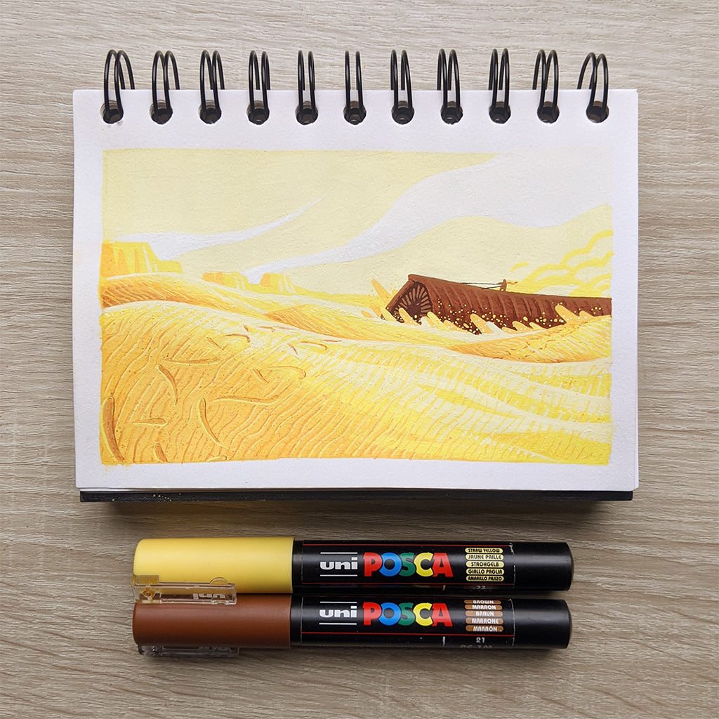 A James Chapman notebook displays artwork inspired by Dune: Part Two, featuring a yellow desert with rolling hills and a distant structure, accompanied by yellow and brown markers on a wooden surface.