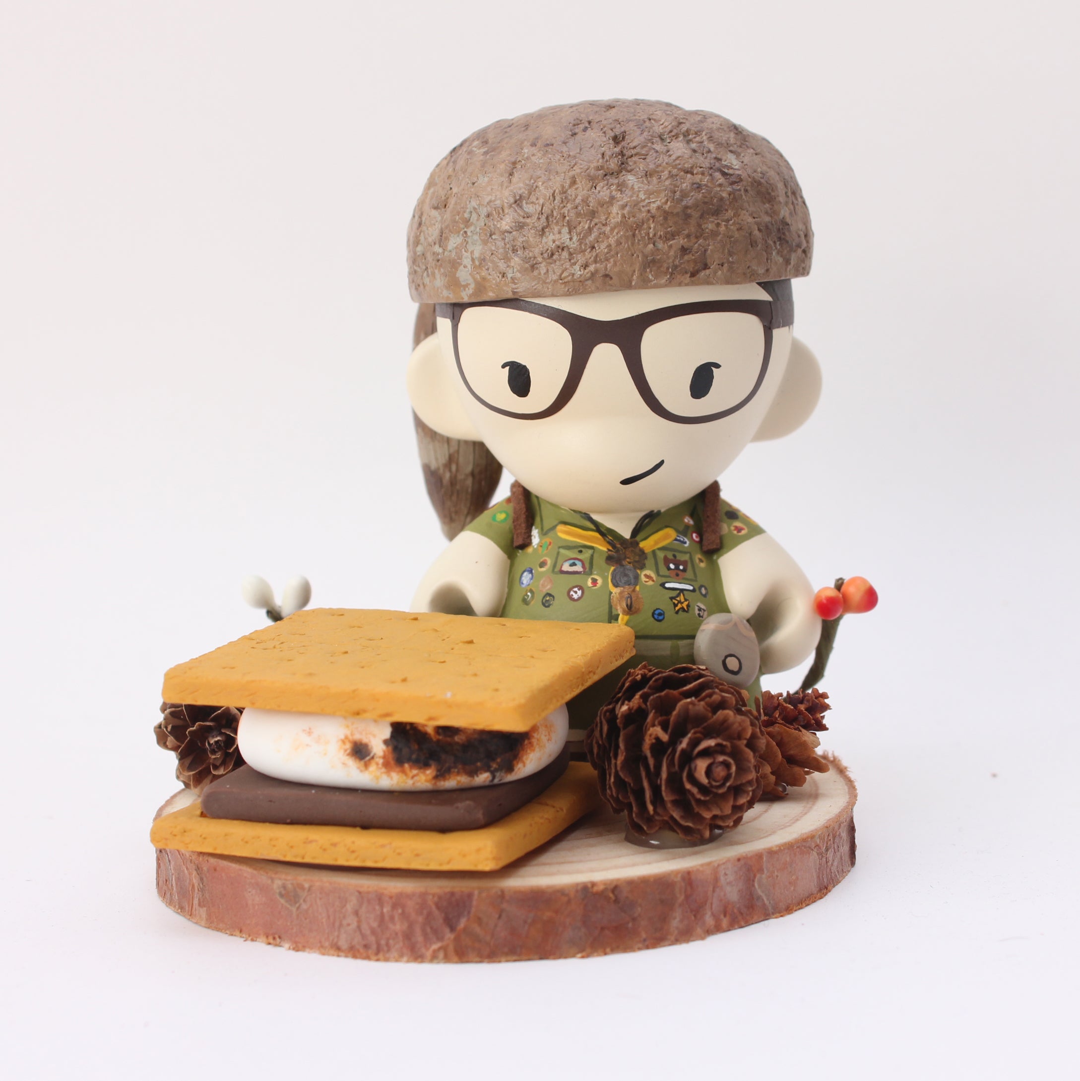 A whimsical vinyl figure by Zard Apuya wears glasses and a nature-themed shirt, holds a small camera, and stands on a wooden base. Part of the Bad Dads IX series, its surrounded by mixed media elements like a crafted smore, pine cones, and a tiny mushroom decoration.