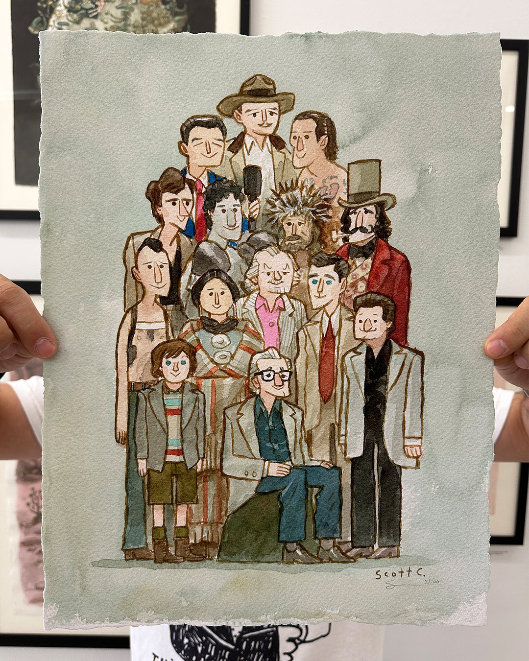 A person holds up a limited edition Scott C. drawing featuring uniquely dressed characters with distinctive hairstyles, standing and sitting together against a light green background. The artwork is hand-signed by the artist.