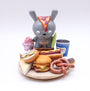 A whimsical mixed media art piece by Spoke Art: a gray bunny toy with a pizza-slice ear stands on a wooden plate, surrounded by toy foods—pretzel, hot dog, burger, sandwich, cheesecake, pink ice cream, and a blue takeout cup.