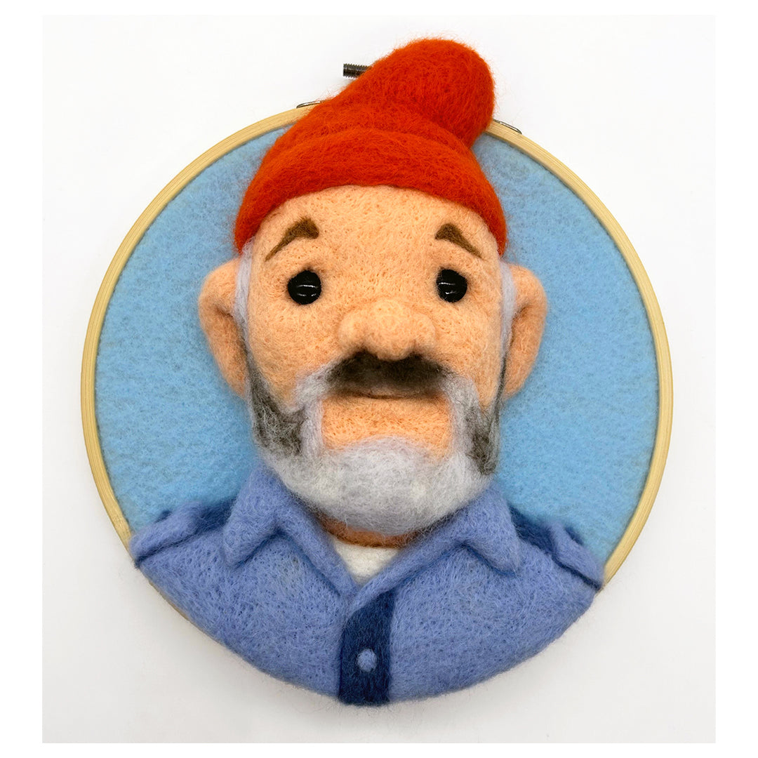 An Ed Mironiuk felt artwork in an embroidery hoop depicts a mustached and bearded person wearing a red beanie and blue shirt on a light blue background, channeling the quirky charm of The Life Aquatic.