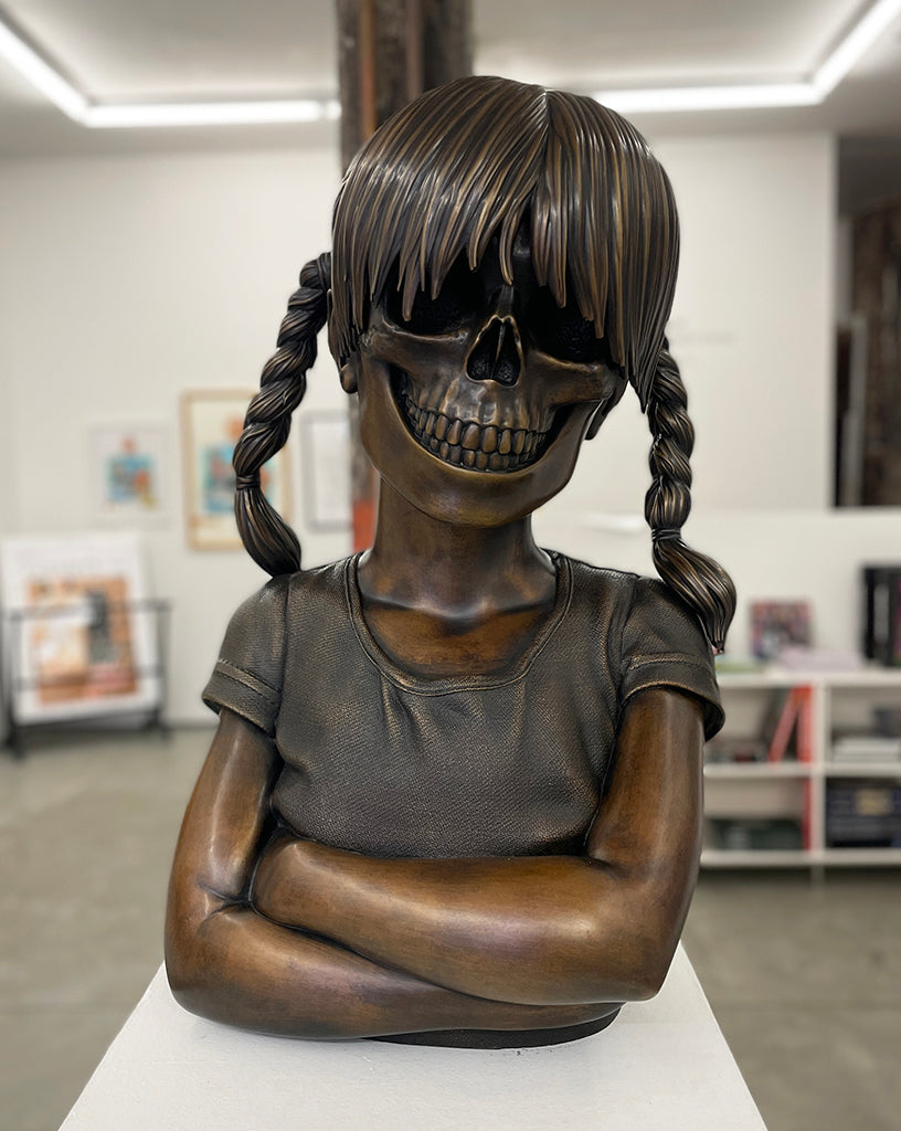 A cast bronze sculpture by Zoltron, titled "Sue Nami Transfiguration," features a girl with braided hair, arms crossed, and a skull in place of her face. Presented by Spoke Art, it's displayed in a modern gallery with framed art and bright lighting.