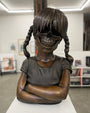 A cast bronze sculpture by Zoltron, titled "Sue Nami Transfiguration," features a girl with braided hair, arms crossed, and a skull in place of her face. Presented by Spoke Art, it's displayed in a modern gallery with framed art and bright lighting.
