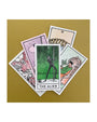 A set of five tarot cards by Brighton Ballard, inspired by a Wes Anderson color palette, lies on a mustard surface. The Alien card is centered in green amidst vibrant designs. This Risograph-printed collection showcases a visual symphony of pastels and bold hues.