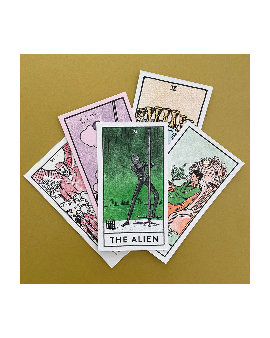 A set of five tarot cards by Brighton Ballard, inspired by a Wes Anderson color palette, lies on a mustard surface. The Alien card is centered in green amidst vibrant designs. This Risograph-printed collection showcases a visual symphony of pastels and bold hues.