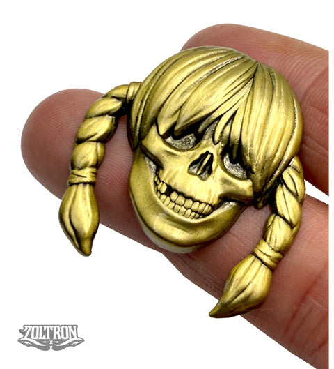 A brass-colored skull pin by ZOLTRON with a wide grin, braided pigtails, bangs, and the VOLTRON logo in the bottom left—this limited edition piece is true Zoltron art.