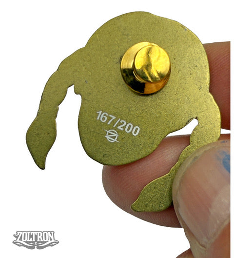 A hand holds a gold-colored ZOLTRON limited edition pin, shaped like a stylized head with long hair. The back displays a clasp, stamped 167/200, with the ZOLTRON logo above an art watermark.