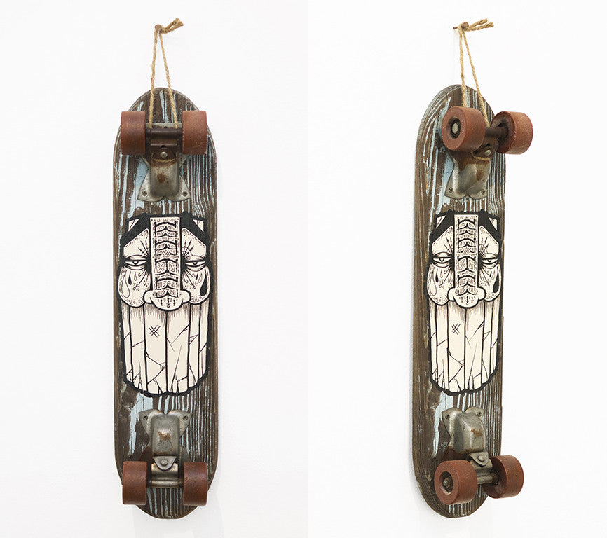 A GATS acrylic skateboard with a tribal-style symmetrical face design in black and white is mounted vertically on the wall. Featuring a vintage look and brown wheels, it was once a highlight at Art Market San Francisco.