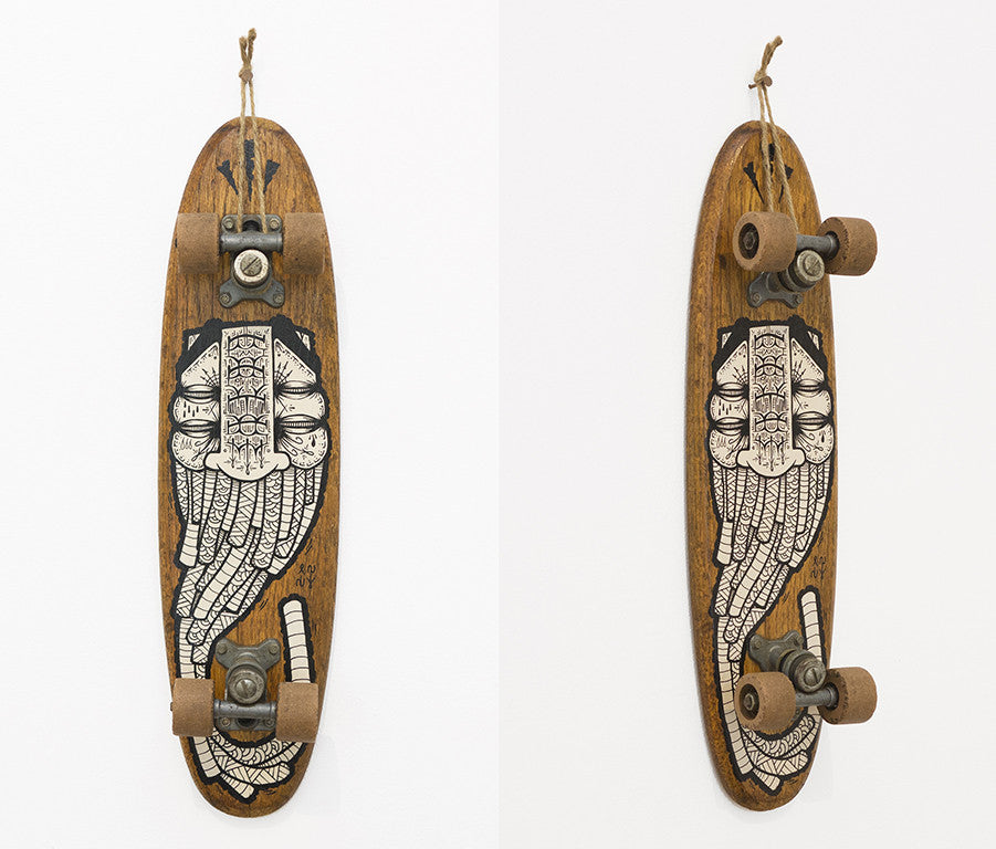 At Art Market San Francisco, a GATS skateboard with a wood finish hangs vertically like art, showcasing a black and white illustration of a snarling wolfs face. The wheels and trucks are visible, with a string attached for exhibition purposes.