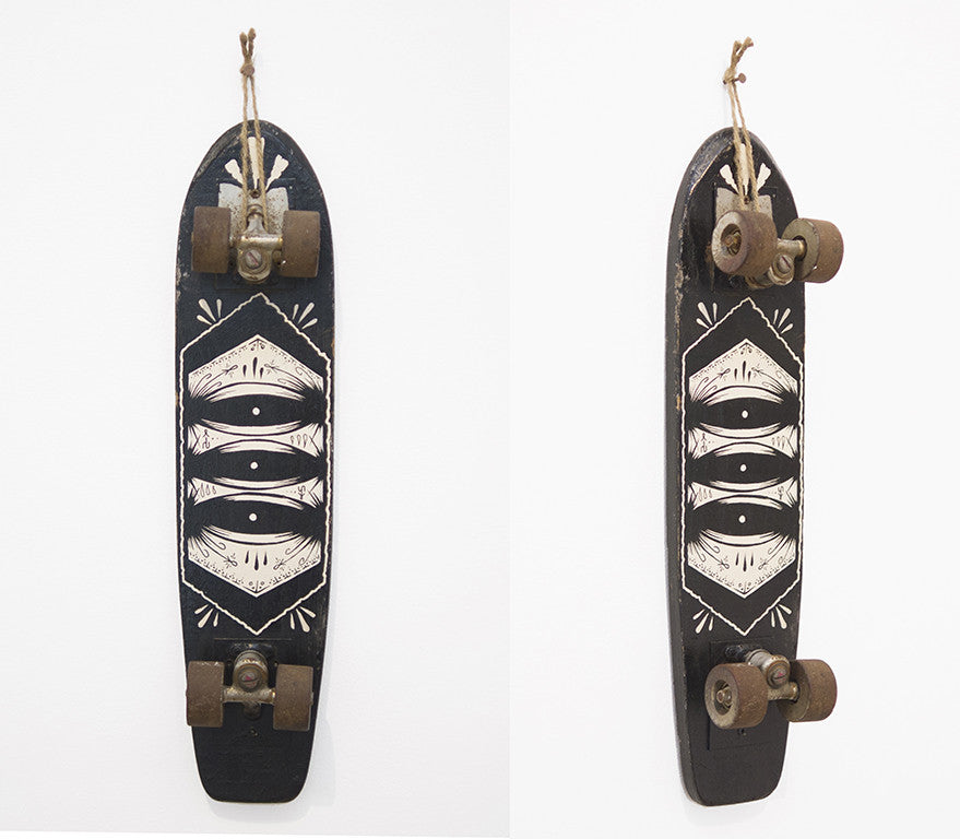 A vintage GATS skateboard features a black deck with an abstract white geometric design, like acrylic art. Wooden and worn, its scuffed brown wheels hang vertically on a white wall, capturing the eclectic essence of Art Market San Francisco.