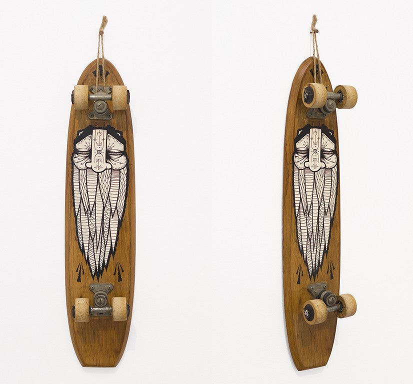 A vintage skateboard featuring GATS black acrylic design of a stylized face with closed eyes is displayed on a white background. With its carved wooden deck, the skateboard hangs upright by a rope, showcasing its trucks and wheels.