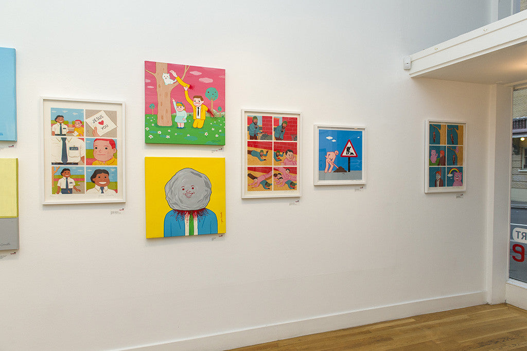 A gallery wall displays custom-framed illustrations in vibrant colors reminiscent of Joan Cornellàs cartoon-style art, set against colorful backgrounds. Below, a wooden floor complements the scene, while a window to the right offers a partial view of a bustling street.