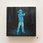 Casey Weldons acrylic painting on wood shows a figure in a cowboy hat and checkered shirt aiming a gun in a forest. The artwork uses blue and black shades for a night-time feel, with a red tag displaying the number 18 beneath this captivating piece for sale.