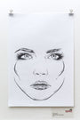 A black and white line drawing of a womans face, reminiscent of a Rhys Cooper sketch, is centered on a white sheet. The focus is on her detailed eyes and lips. This ink-on-paper artwork is clipped at the top and displayed on a pristine white wall.