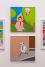 Two colorful paintings by Joan Cornellà hang on a wall. The top features a person in a suit with flaming hair and a smiling figure near a tree, while the bottom shows Untitled #12 where someone with extended legs walks in their hat and suit.