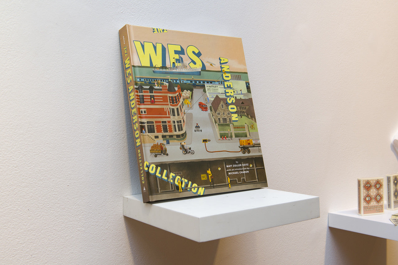The image features the book The Wes Anderson Collection by Matt Zoller Seitz, displaying Wes Anderson’s filmography on a small white shelf against a light-colored wall. The cover showcases colorful building and vehicle illustrations, highlighting Wes Andersons whimsical world.