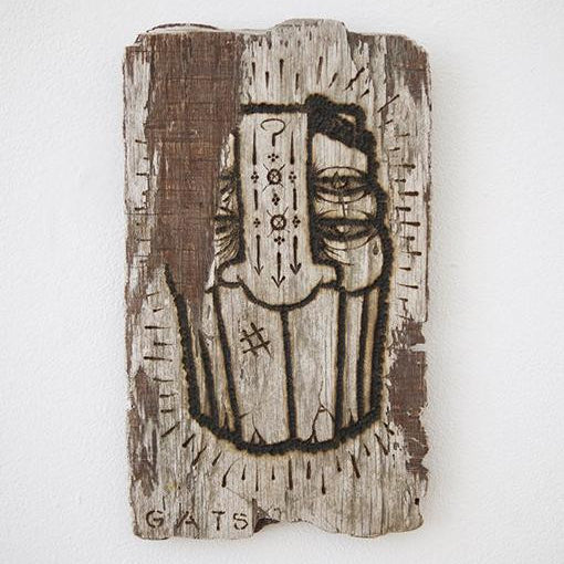A rustic wooden artwork by GATS, carved from burnt driftwood featuring a face with eyes and symbolic patterns such as arrows and a question mark. It measures 6.25 x 10.5 and is signed GATS at the bottom, showcasing a textured, aged appearance.