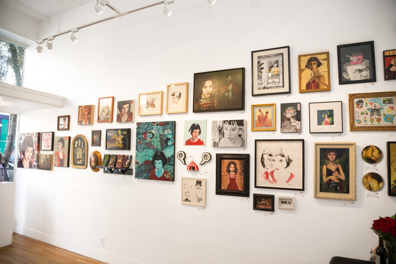 A gallery wall displays a diverse collection of framed artworks, featuring portraits and acrylic panel pieces by Kelsey Beckett. Various sizes and styles are arranged on a white wall, with a ceiling light highlighting the vibrant and eclectic Sur Le Fil display.