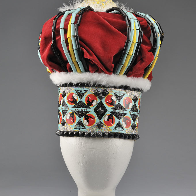 A vibrant Lucien Shapiro Crown II, displayed on a white stand, features a red velvet dome with blue, black, and multicolored geometric patterns. Trimmed with fur and topped by a golden bejeweled finial, this mixed media masterpiece is captivating.