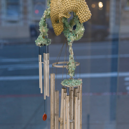 Lucien Shapiros abstract sculpture, with textured yellow and green organic shapes on metal rods and chimes, hangs in front of a window. The background reveals a blurred street scene through the glass, capturing the vibe of the Supersonic Invitational.