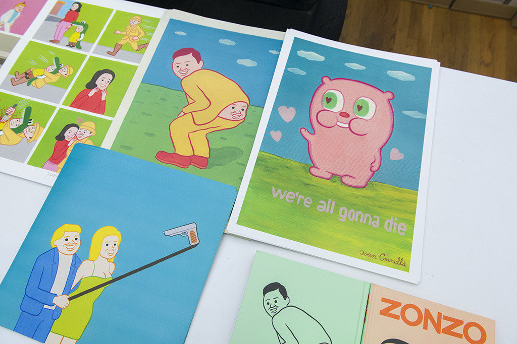 A table showcases vibrant posters and books. One digital print features a pink creature with Gonnadie text, while another highlights Joan Cornellàs iconic style with a person in a yellow suit with their face on their backside. Nearby are illustrated covers, including one titled Zonzo.