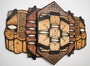 A geometric wooden wall sculpture by GATS features stylized symmetrical faces in an abstract design. Crafted with enamel on wood, it showcases intricate patterns and shapes in earthy tones of brown and beige.