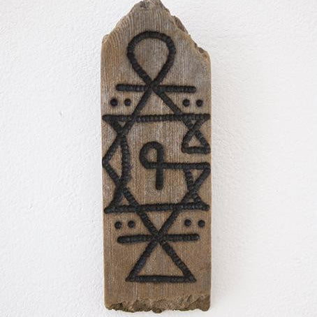This GATS wooden plaque features carved geometric patterns—triangles, circles, and a top curved loop. Its central area showcases a stylized P. Crafted from burnt driftwood for a rustic look, its mounted on a white wall.