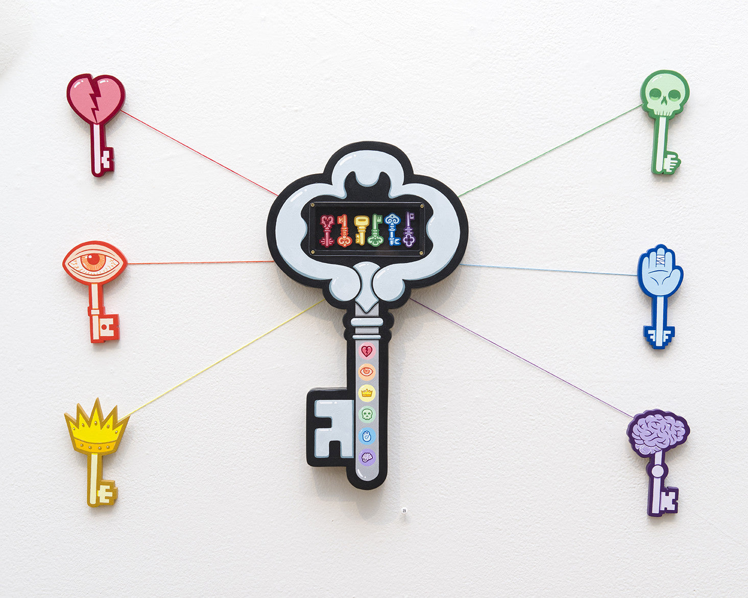 Matt Ritchies wall art features a central key with a digital display, surrounded by vibrant icons. Made from acrylic on wood, the set includes six smaller keys—a heart, skull, hand, brain, crown, and eye—each uniquely colored and connected by lines.