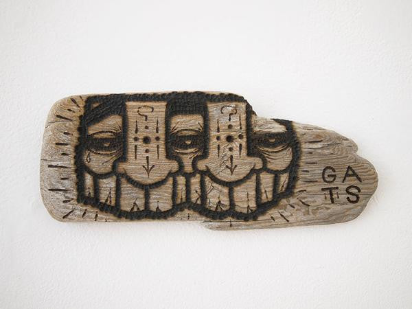 A wooden piece of burnt driftwood with carved, stylized three-eyed faces and intricate linework features the inscription GATS on the right side. This striking wood burn art is influenced by urban and tribal motifs.