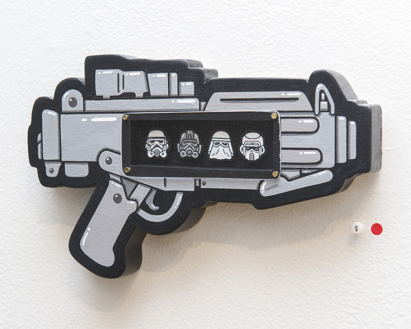 A stylized gun-shaped wall art by Matt Ritchie, crafted from hand-cut wood in black, gray, and white. It displays three cartoon character faces inspired by Storm Troopers within a rectangle cutout, plus a small red dot sticker on the neighboring white wall.