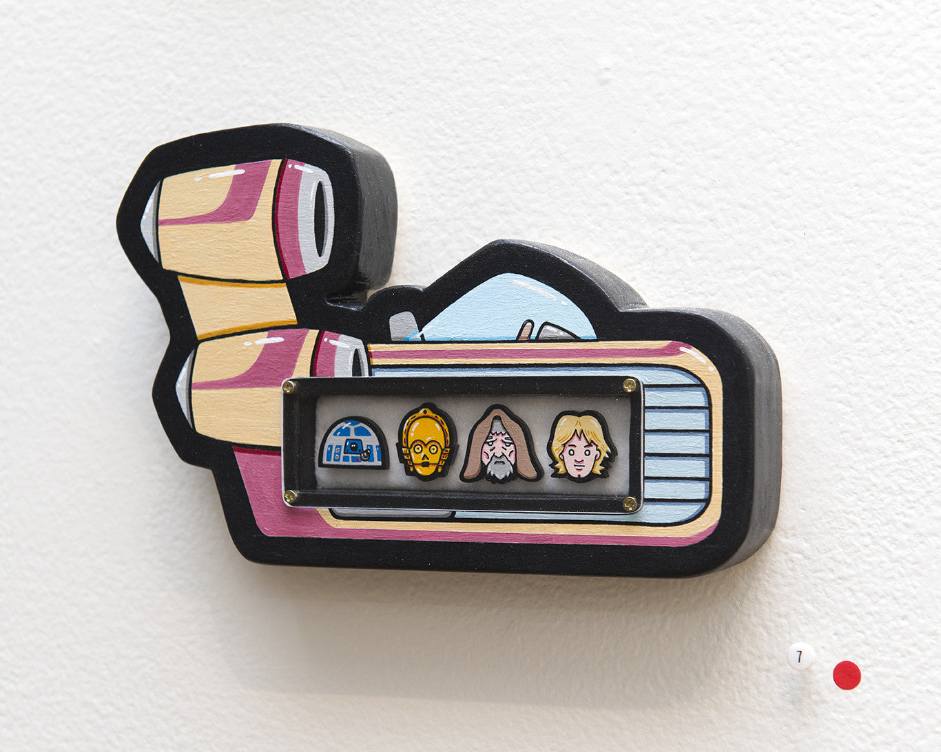 A Matt Ritchie wooden artwork, hand-cut with precision in the shape of a film camera, features cartoon icons of a house, yellow robot head, bearded man, and blonde person. This acrylic piece is mounted on a white wall with a small red dot beside it.