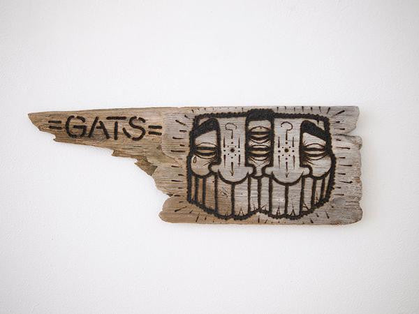 A weathered driftwood piece features a stylized painting of two faces with elongated eyes and neutral expressions, with GATS expertly burnt into the left side, blending rustic charm and urban flair.