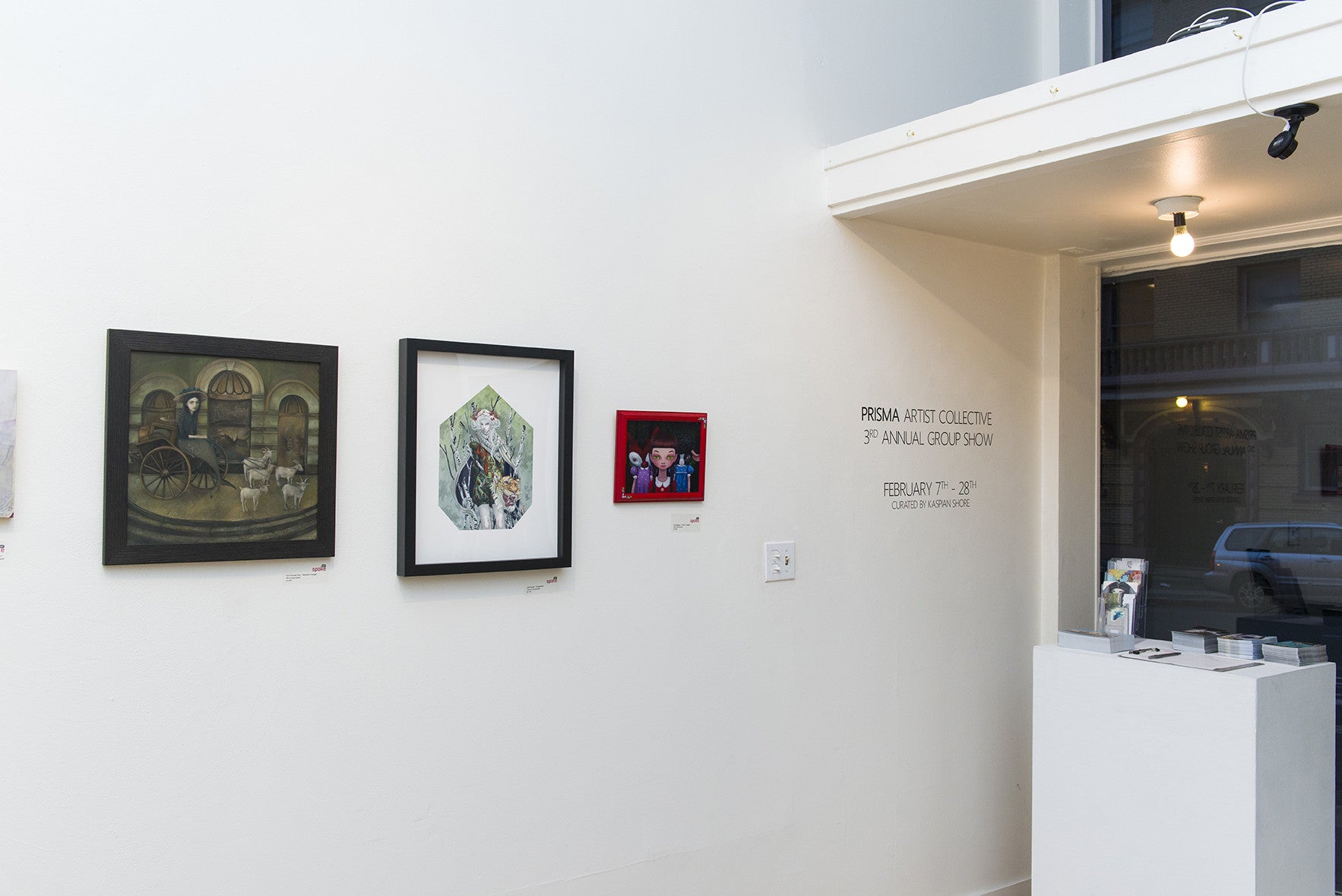 The gallery wall displays three framed artworks: a cart scene, an abstract design, and a vibrant portrait of Trick or Treaters. The text reads Prisma Artist Collective 3rd Annual Group Show February 27-28th Curated by Kelly Vivanco, featuring works like those from Ana Bagayan.