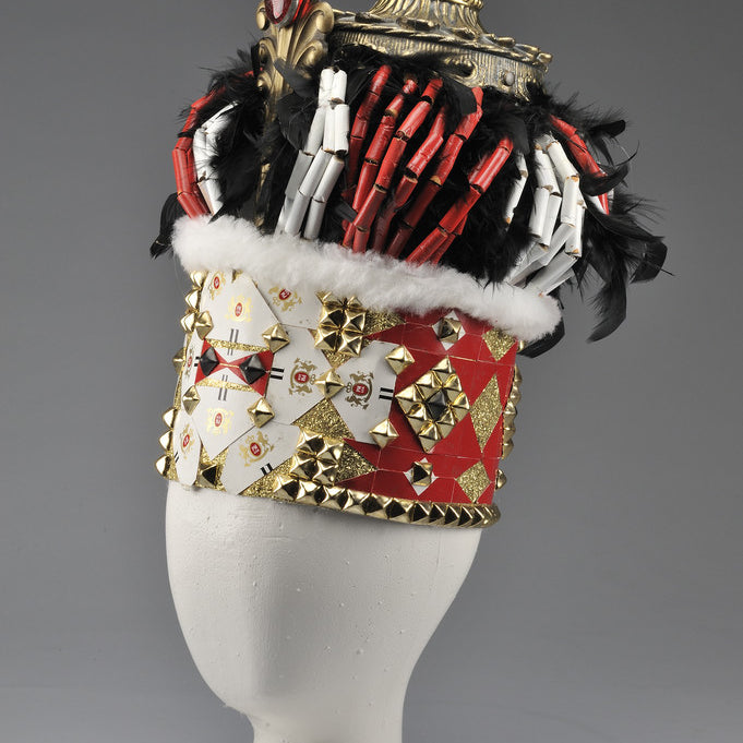 In Lucien Shapiros Crown III collection, a tall, ornate hat features a gold top, red and white dice, black feathers, a red velvet band with gold studs, and white fur trim—an intricate design embracing a playing card and dice theme.