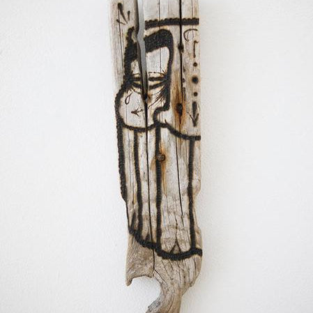 A piece of burnt driftwood from GATS stands vertically, showcasing abstract black ink drawings of a stylized face and body. The charred surface creates a striking contrast against the plain white background.