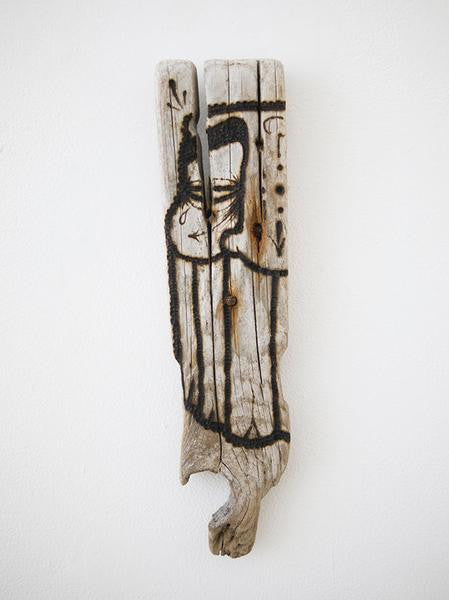 A piece of burnt driftwood from GATS stands vertically, showcasing abstract black ink drawings of a stylized face and body. The charred surface creates a striking contrast against the plain white background.