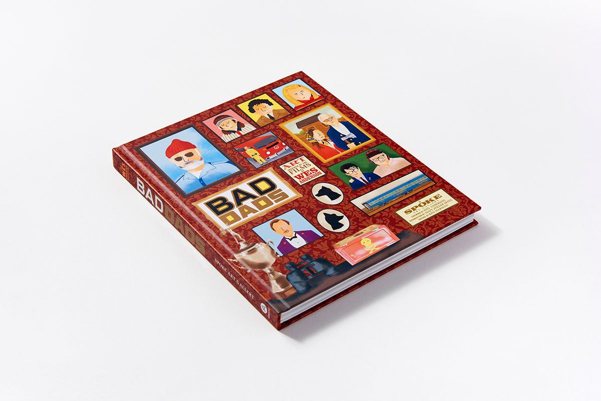 The image shows the book Bad Dads: Art Inspired by Wes Anderson from Matt Zoller Seitz, a tribute to the Bad Dads exhibition at Spoke Art Gallery. Its cover has minimalist illustrations and scene collages on a red patterned background, with the book placed on a white surface.