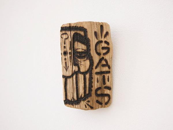A small piece of burnt driftwood with abstract facial features and the word GATS carved and painted on it is mounted on a plain white wall.