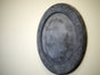 A round, metallic shield-like piece by Andrew Witrak is displayed at Spoke Art Gallery. It hangs on a plain wall and showcases an etched geometric border with a weathered, textured finish similar to Witraks pool light installations.