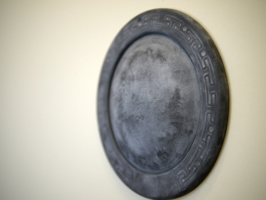 A round, metallic shield-like piece by Andrew Witrak is displayed at Spoke Art Gallery. It hangs on a plain wall and showcases an etched geometric border with a weathered, textured finish similar to Witraks pool light installations.