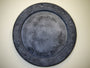 A round, weathered concrete manhole cover by Andrew Witrak features a decorative border with a geometric pattern, reminiscent of Spoke Art Gallerys style. Its textured surface and light background evoke the look of pool light reflections.