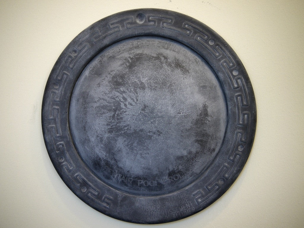 A round, weathered concrete manhole cover by Andrew Witrak features a decorative border with a geometric pattern, reminiscent of Spoke Art Gallerys style. Its textured surface and light background evoke the look of pool light reflections.