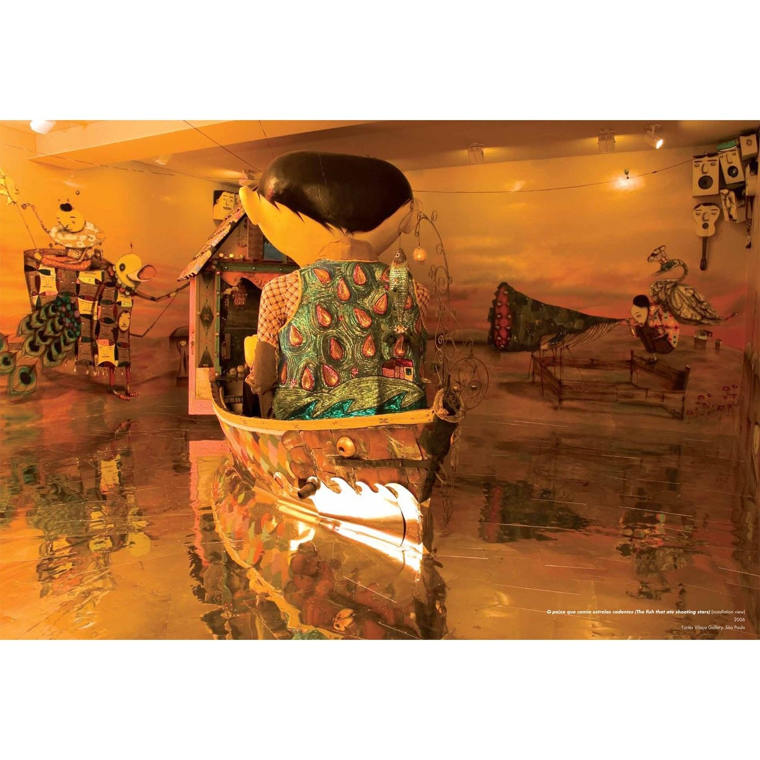 A surreal Os Gemeos-inspired art installation showcases a large head figure seated in a boat on reflective water, surrounded by whimsical, colorful characters and abstract graffiti shapes, all within a warmly golden-lit room.